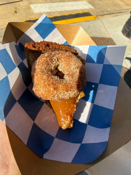 A cinnamon-sugar cider donut sitting on a slice of crumb-topped apple pie in a blue-checked paper boat