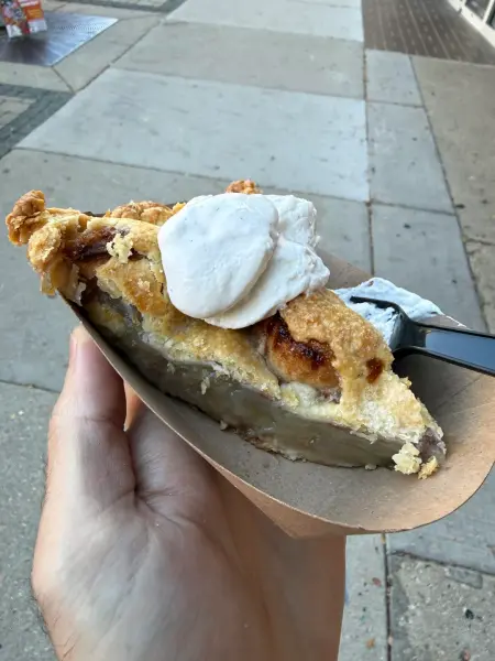 A hand holding a paper boat with a slice of double-crust apple pie topped with whipped cream and a black plastic fork